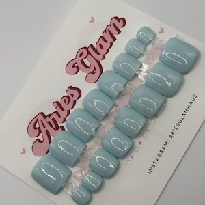 May include: A set of light blue press-on toenails in different sizes, presented on a white card with pink text reading "Aries Glam". The nails have a glossy finish and a square shape. The card also shows the Instagram handle "ariesglamhaus".