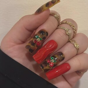 May include: A hand with long, square-shaped acrylic nails. Some nails feature a tortoiseshell pattern with cherry accents, while others are solid red. Gold rings adorn the fingers. The nails are a bold fashion statement.
