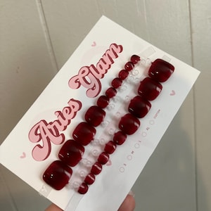 Puede incluir: Un juego de uñas postizas para los dedos de los pies en un tono rojo oscuro, dispuestas sobre una tarjeta blanca con el texto "Aries Glam" en una fuente retro. Las uñas son de varios tamaños y están listas para su aplicación.