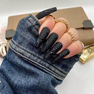May include: Matte black long coffin-shaped artificial nails with glossy black drips. The hand is adorned with multiple gold rings. The nails are set against a denim jacket and a tan leather wallet.