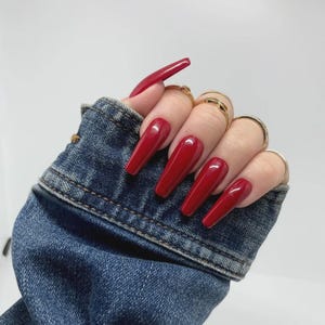 Op de afbeelding: Een hand met lange, kistvormige, rode acrylnagels. De nagels zijn diep, glanzend rood. De hand is versierd met meerdere gouden ringen en wordt gedeeltelijk bedekt door een blauw spijkerjasje.