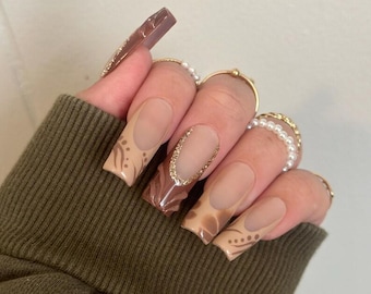 May include: Long, square-shaped acrylic nails with a nude base and brown and gold designs. Some nails feature a French tip with gold glitter, while others have brown patterns. Gold rings with pearls and small gold bands adorn the fingers.