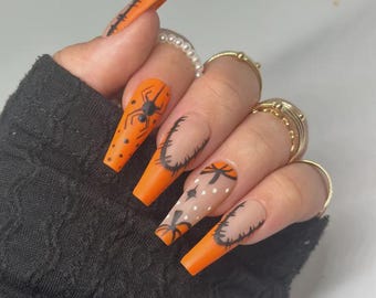 Pode incluir: Unhas postiças longas, laranja e nude em forma de caixão com desenhos pretos com tema de Halloween. Os desenhos incluem aranhas, teias de aranha e um padrão de pontos. As unhas são acentuadas com anéis dourados e um anel de pérolas.