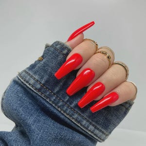 May include: A hand with long, bright red coffin-shaped acrylic nails. The hand is adorned with multiple gold rings. The nails are a vibrant, glossy red, contrasting with the blue denim jacket sleeve in the background.