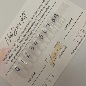 Op de afbeelding: Een nagelmatenkit met doorzichtige, genummerde nageltips op een kaart. De kaart is voorzien van de tekst "Nail Sizing Kit" en instructies. De kit helpt klanten bij het bepalen van hun nagelmaten voor persnagels.