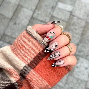 May include: A hand with long, pointed fingernails featuring various designs. Some nails have a black and white checkered pattern, while others showcase cherries, stars, and leopard print. Gold rings adorn the fingers, and the hand is partially covered by a plaid sleeve.