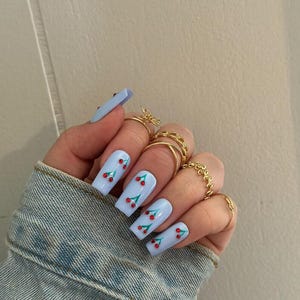 May include: Light blue, square-shaped artificial nails with a cherry design. Each nail features a light blue base with a pattern of red cherries and green leaves. The hand also has multiple gold rings and is wearing a denim jacket.