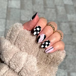 May include: A hand with a manicure featuring a variety of designs. The nails showcase a mix of black and white checkered patterns, pink and black swirls, and solid black and pink nails. Gold rings adorn the fingers.