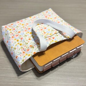 May include: A floral print tote bag with a white handle holds a tan book with labeled tabs. The bag features a white background with a pattern of small flowers in shades of pink, yellow, and blue. The book is partially visible.