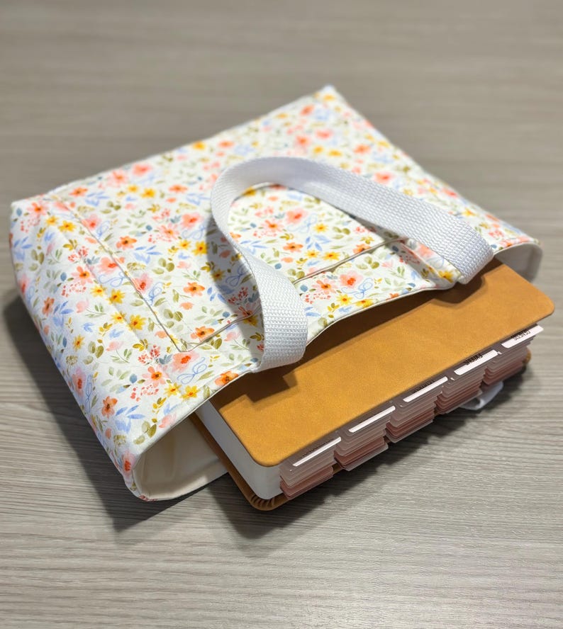 May include: A floral print tote bag with a white handle holds a tan book with labeled tabs. The bag features a white background with a pattern of small flowers in shades of pink, yellow, and blue. The book is partially visible.