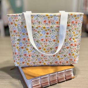 May include: A floral print tote bag with white handles and a front pocket, resting on a large book with labeled tabs. The bag features a pattern of pink, yellow, and blue flowers on a white background.