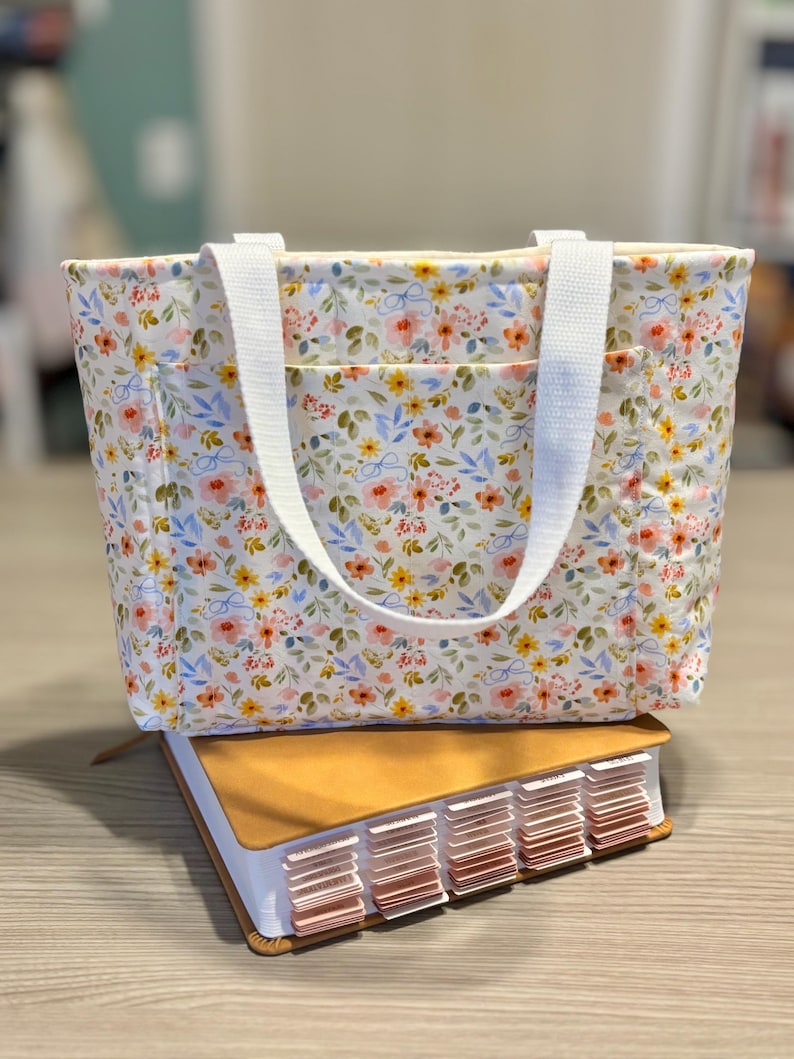 May include: A floral print tote bag with white handles and a front pocket, resting on a large book with labeled tabs. The bag features a pattern of pink, yellow, and blue flowers on a white background.