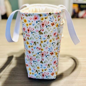 May include: A floral print tote bag with white handles. The bag features a white background with a pattern of colorful flowers in shades of pink, yellow, and blue. The interior is a solid cream color.