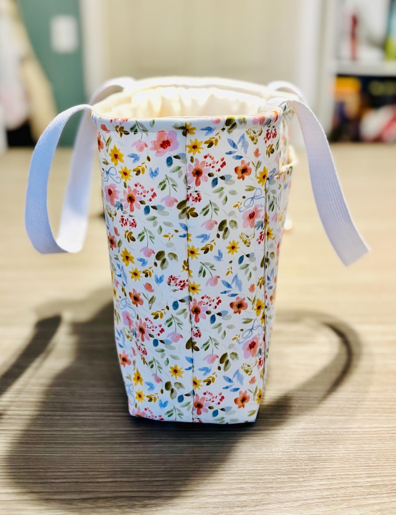 May include: A floral print tote bag with white handles. The bag features a white background with a pattern of colorful flowers in shades of pink, yellow, and blue. The interior is a solid cream color.