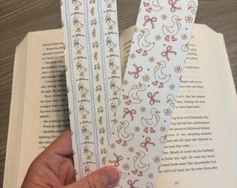 May include: Two decorative bookmarks with duck and bow patterns. One bookmark has a vertical striped pattern with ducks, bows, and flowers. The other bookmark has a repeating pattern of ducks and bows. The bookmarks are held over an open book.