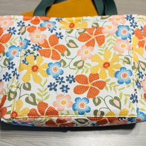 May include: A floral tote bag with a white background and a pattern of orange, yellow, and blue flowers. The bag has a green handle and a pocket on the front. A yellow item is visible inside the bag.