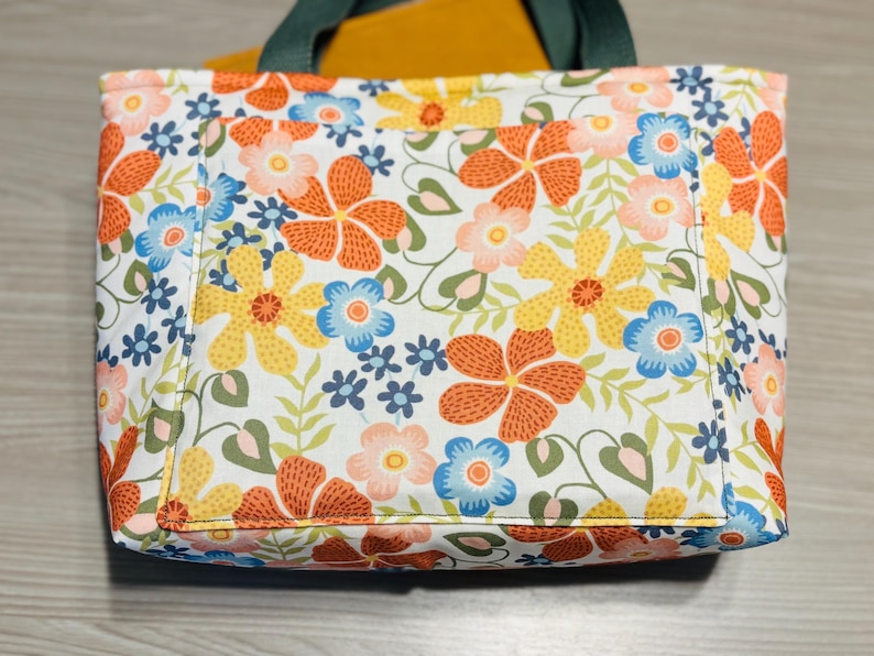 May include: A floral tote bag with a white background and a pattern of orange, yellow, and blue flowers. The bag has a green handle and a pocket on the front. A yellow item is visible inside the bag.