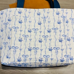 May include: A rectangular tote bag with a white background and a blue floral and ribbon pattern. The bag has a blue handle and a pocket on the front. The interior is orange.