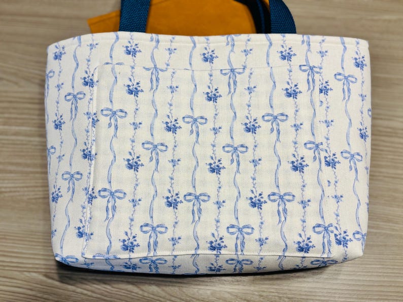 May include: A rectangular tote bag with a white background and a blue floral and ribbon pattern. The bag has a blue handle and a pocket on the front. The interior is orange.