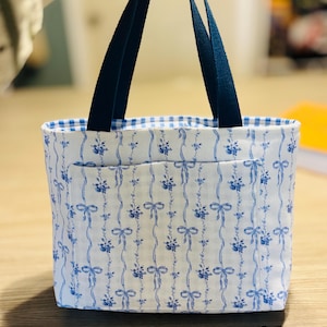 May include: A white tote bag with a blue floral and ribbon pattern. The bag has a blue gingham lining and navy blue handles. The bag has a front pocket and is made of fabric.