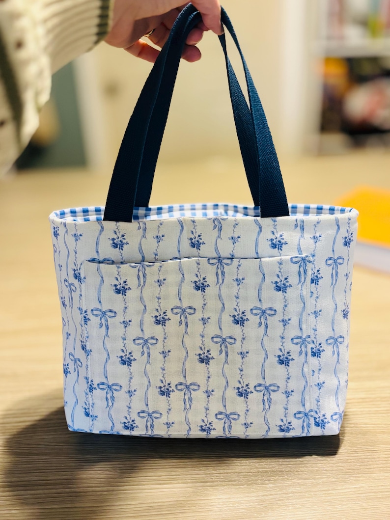 May include: A white tote bag with a blue floral and ribbon pattern. The bag has a blue gingham lining and navy blue handles. The bag has a front pocket and is made of fabric.