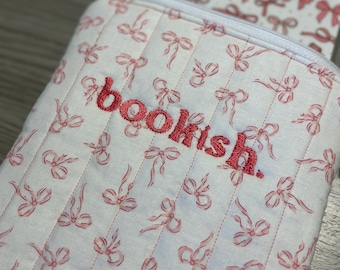 May include: A white quilted case with a repeating pattern of pink bows. The word "bookish" is embroidered in red on the front. A matching item is visible in the background, also with a bow pattern.