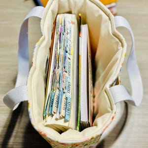 May include: A floral print tote bag with white handles, filled with books and notebooks. The bag's interior is a cream color, and the books have colorful tabs. The bag is sitting on a wooden surface.