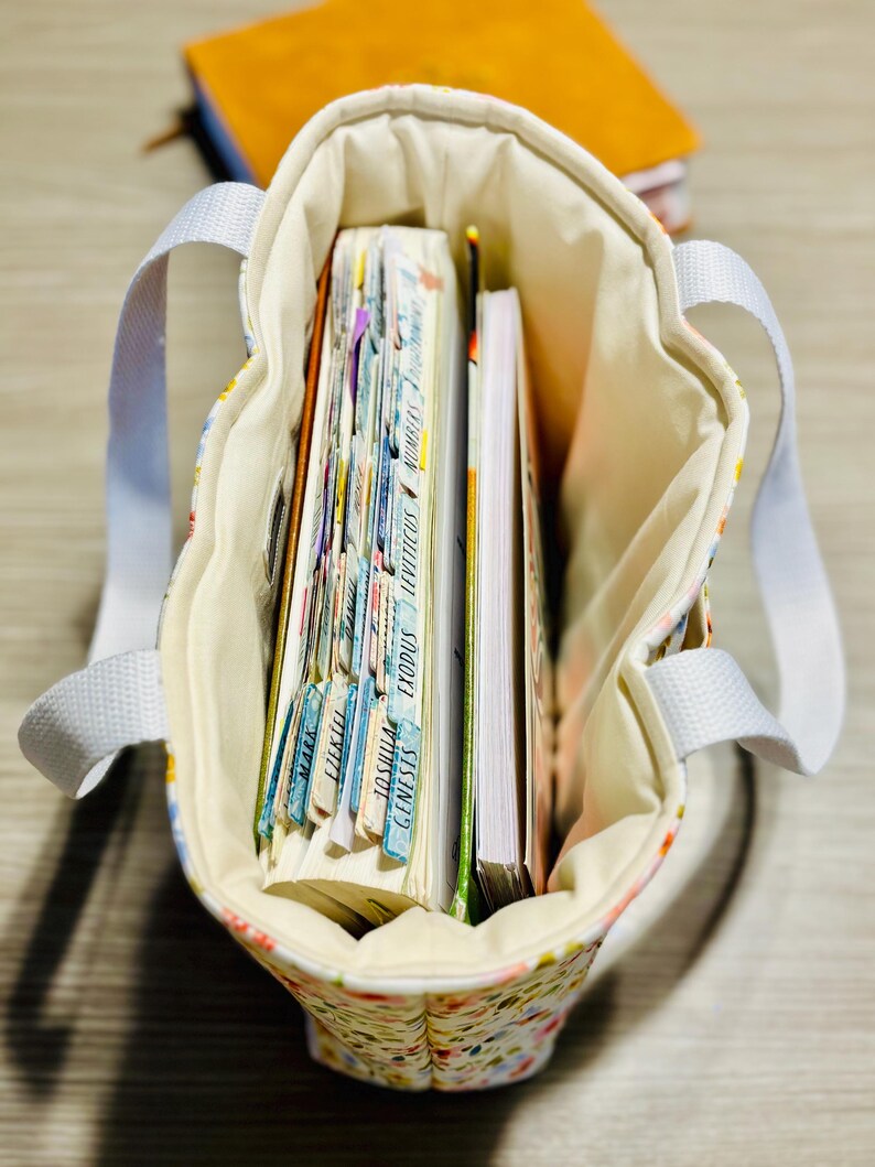 May include: A floral print tote bag with white handles, filled with books and notebooks. The bag's interior is a cream color, and the books have colorful tabs. The bag is sitting on a wooden surface.