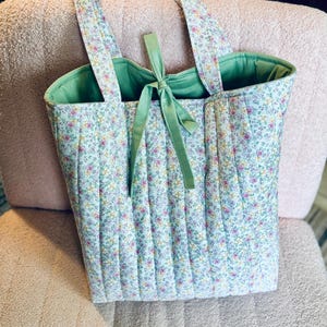 May include: A quilted tote bag with a floral print in shades of pink, yellow, and green. The bag has a green lining and a green ribbon tied at the top.