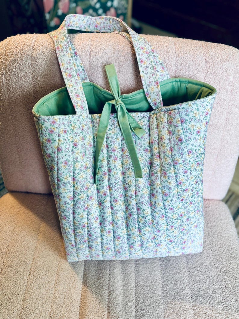 May include: A quilted tote bag with a floral print in shades of pink, yellow, and green. The bag has a green lining and a green ribbon tied at the top.