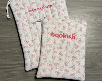 May include: Two white quilted book sleeves with a pink bow pattern. One sleeve has the text "one more chapter" embroidered in pink, and the other says "bookish."