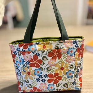May include: A floral tote bag with a white background and a pattern of red, yellow, and blue flowers. The bag has dark green handles and a green interior lining. The bag is held up by a person.