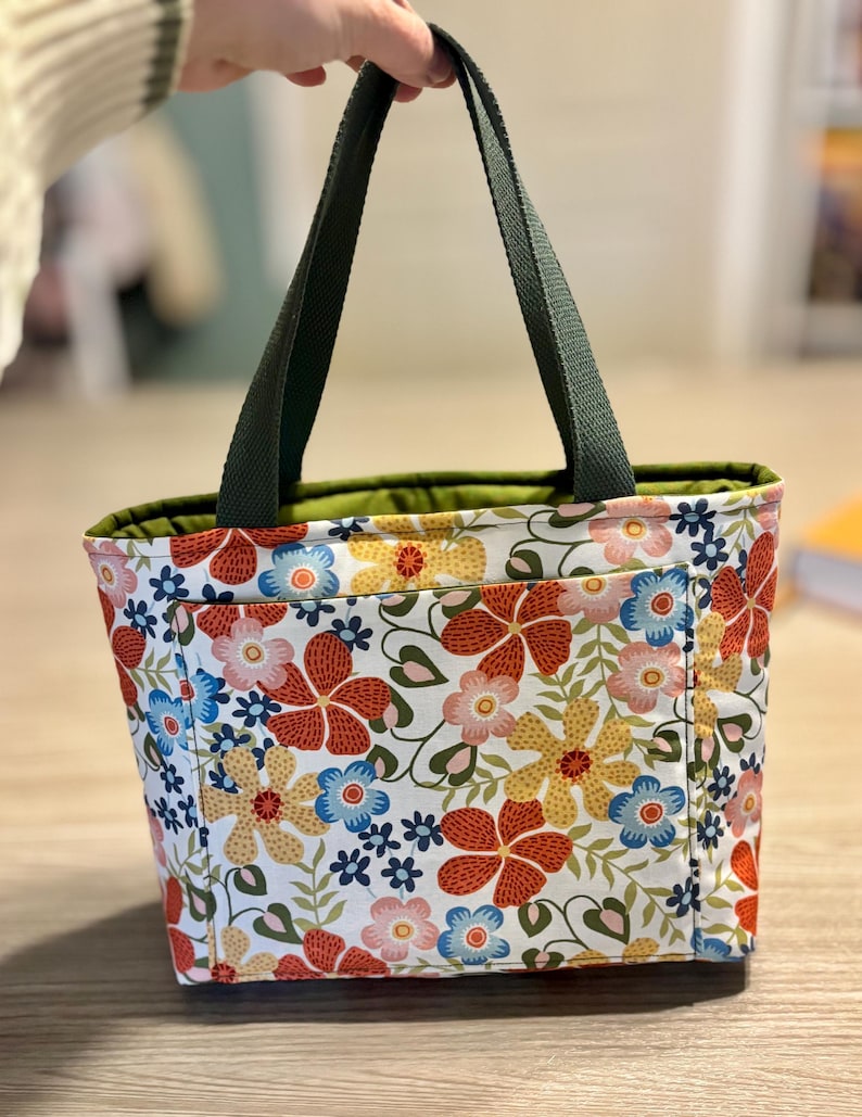 May include: A floral tote bag with a white background and a pattern of red, yellow, and blue flowers. The bag has dark green handles and a green interior lining. The bag is held up by a person.