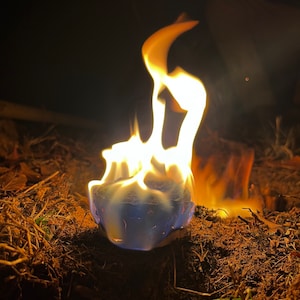 May include: A blue fire starter cube burning with bright orange flames in a dark setting. The cube is surrounded by dry grass and leaves.