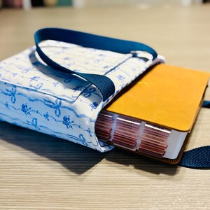 May include: A fabric Bible cover with a blue and white floral pattern, holding a Bible with a tan cover and labeled tabs. The cover has a navy blue handle and strap. The Bible is partially visible.