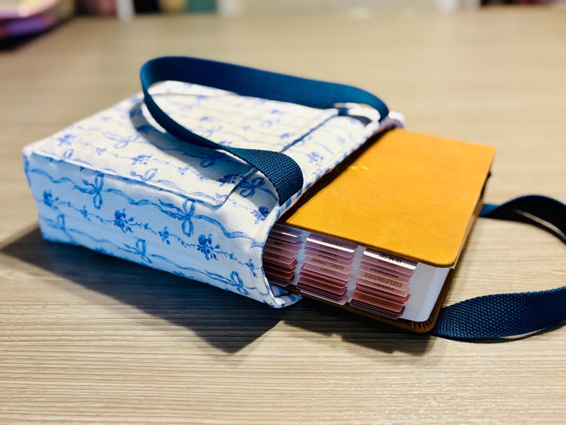 May include: A fabric Bible cover with a blue and white floral pattern, holding a Bible with a tan cover and labeled tabs. The cover has a navy blue handle and strap. The Bible is partially visible.