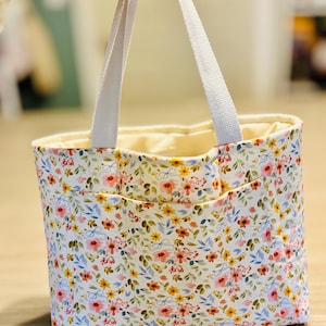 May include: A floral print tote bag with white handles. The bag features a white background with a pattern of pink, yellow, and blue flowers. The interior is a light cream color.