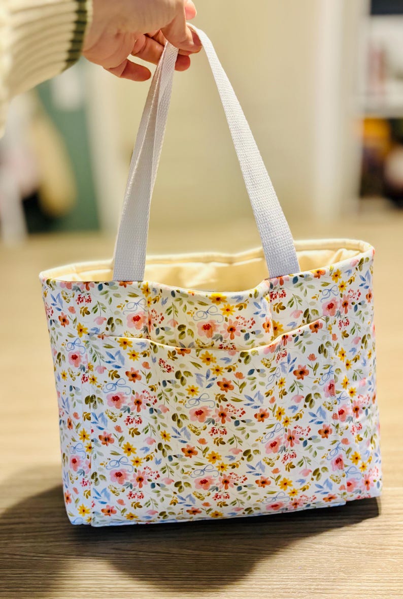 May include: A floral print tote bag with white handles. The bag features a white background with a pattern of pink, yellow, and blue flowers. The interior is a light cream color.