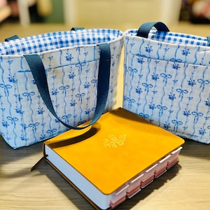 May include: Two small tote bags with blue floral patterns and blue handles. The bags have a gingham lining. A yellow journal with a decorative emblem and page dividers sits in front of the bags.