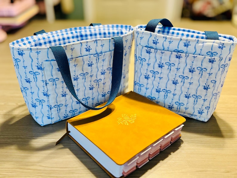 May include: Two small tote bags with blue floral patterns and blue handles. The bags have a gingham lining. A yellow journal with a decorative emblem and page dividers sits in front of the bags.