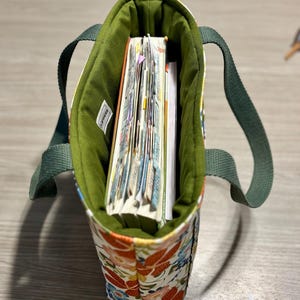 May include: A small tote bag with a floral pattern in orange, blue, and red. The interior is lined with green fabric, and the bag contains several notebooks. The handles are a solid green.