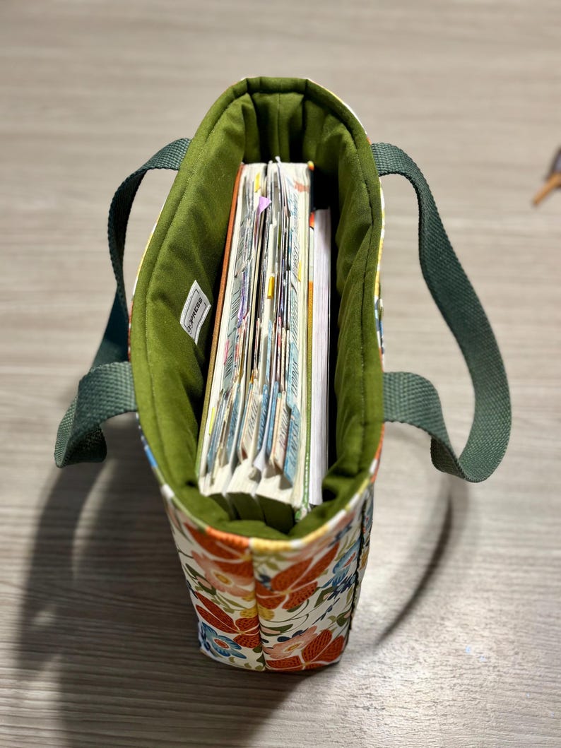 May include: A small tote bag with a floral pattern in orange, blue, and red. The interior is lined with green fabric, and the bag contains several notebooks. The handles are a solid green.