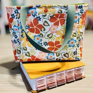 May include: A floral print tote bag with a green handle sits atop a yellow book with labeled tabs. The bag features a white background with red, blue, and yellow flowers. The book has a leather-like cover.