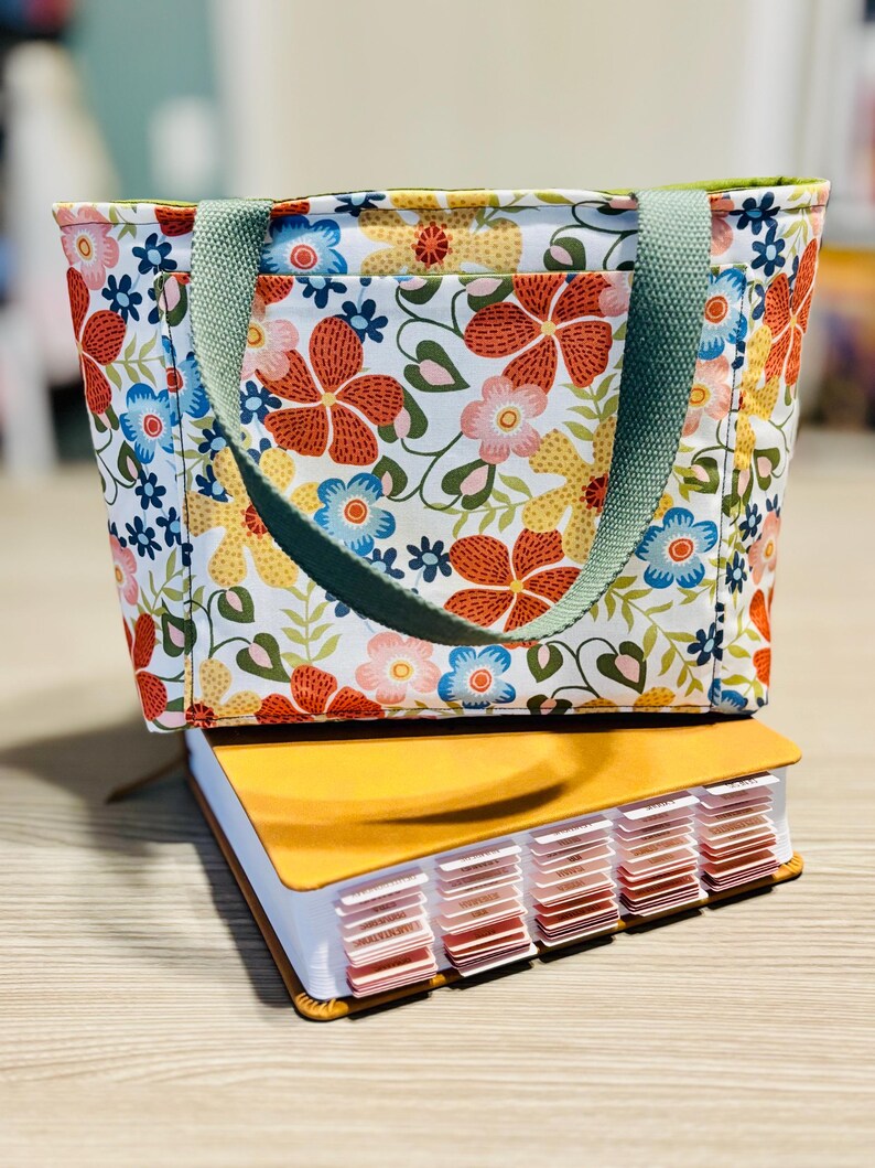 May include: A floral print tote bag with a green handle sits atop a yellow book with labeled tabs. The bag features a white background with red, blue, and yellow flowers. The book has a leather-like cover.