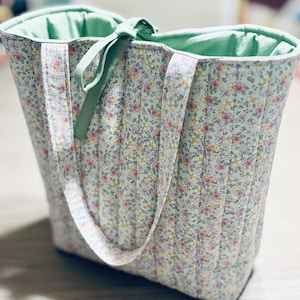 May include: A quilted tote bag with a floral print in shades of pink, yellow, and green. The bag has two green fabric handles and a green fabric lining.