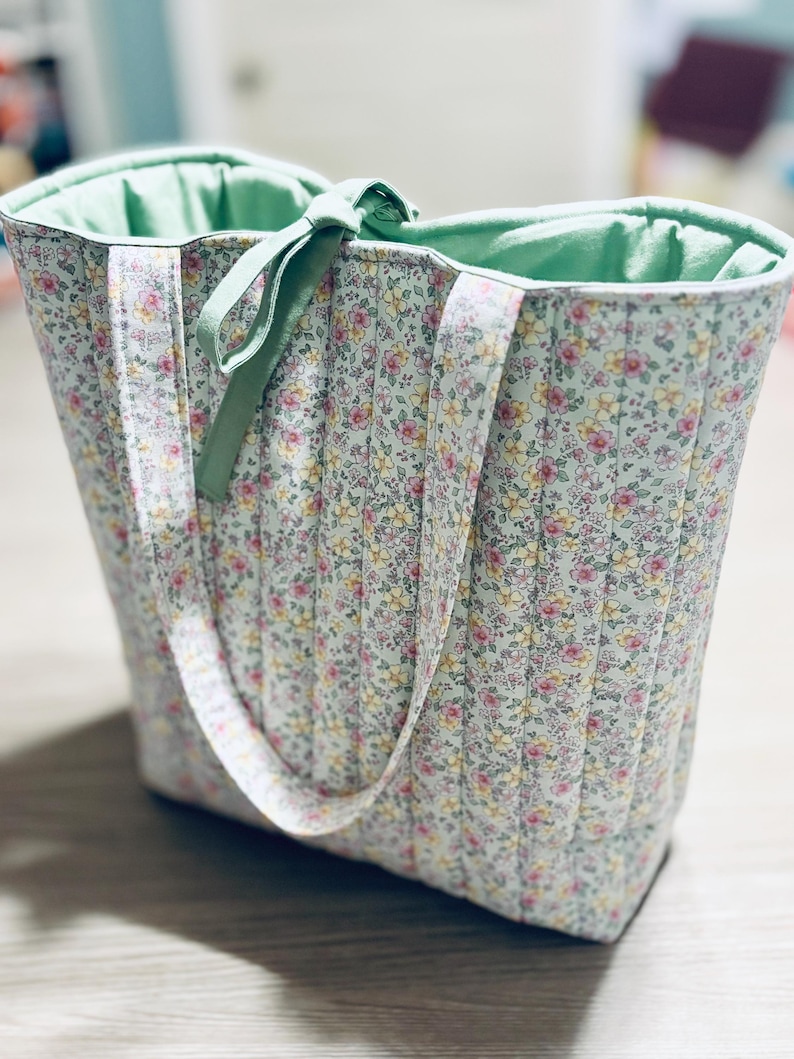 May include: A quilted tote bag with a floral print in shades of pink, yellow, and green. The bag has two green fabric handles and a green fabric lining.