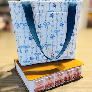 May include: A small tote bag with blue floral and ribbon pattern, blue handles, and a pocket. The bag sits atop a brown leather-bound notebook with labeled tabs. The bag's fabric has a white background.
