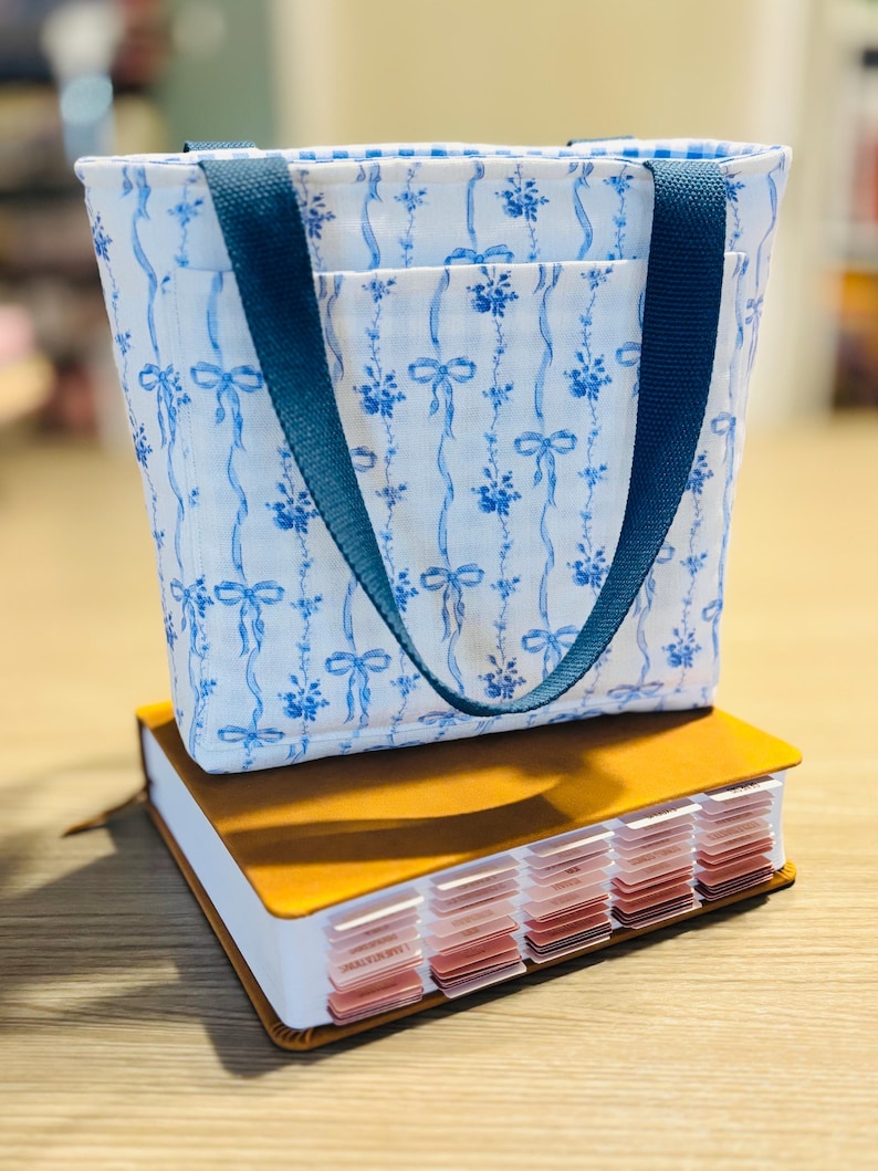 May include: A small tote bag with blue floral and ribbon pattern, blue handles, and a pocket. The bag sits atop a brown leather-bound notebook with labeled tabs. The bag's fabric has a white background.