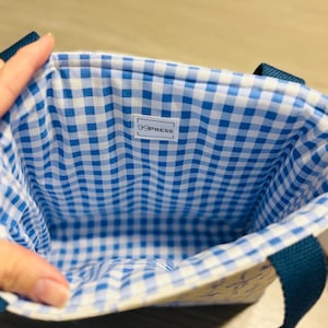 May include: A small tote bag with a blue and white gingham lining. The exterior is a light cream color with a subtle pattern. The bag has dark blue handles and the text "99PRESS" is visible on a white label inside the bag.