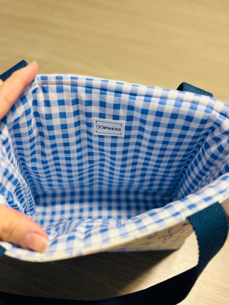 May include: A small tote bag with a blue and white gingham lining. The exterior is a light cream color with a subtle pattern. The bag has dark blue handles and the text "99PRESS" is visible on a white label inside the bag.